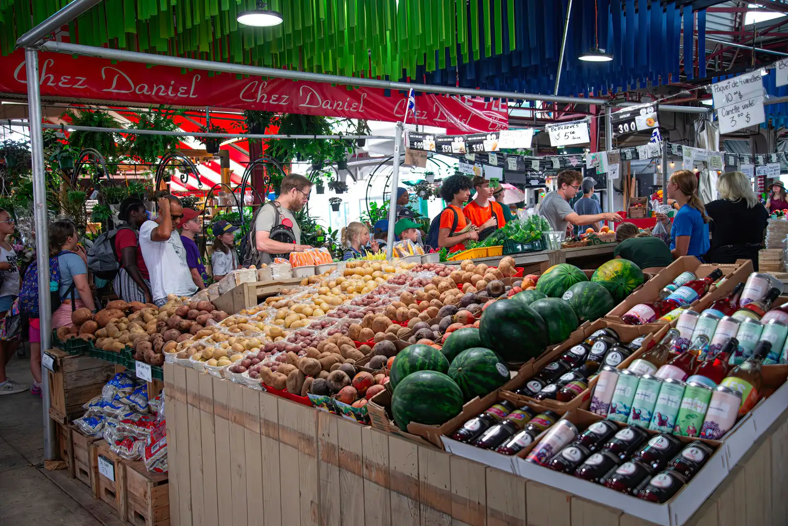 Shoppers at fruit and vegetable stalls under a colourful canopy at a Montreal market